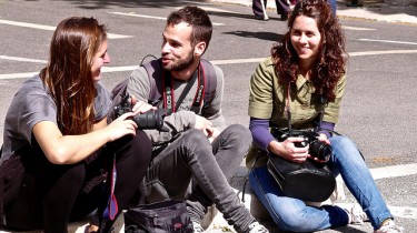 Picture by Pedro Ribeiro Simões. Three photographers in their mid-twenties sitting on the curb, chatting and smiling. A male in the middle of two girls.